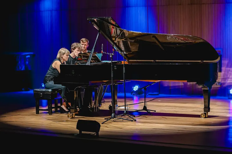 The two pianists side by side at the grand piano on a stage washed in blue light