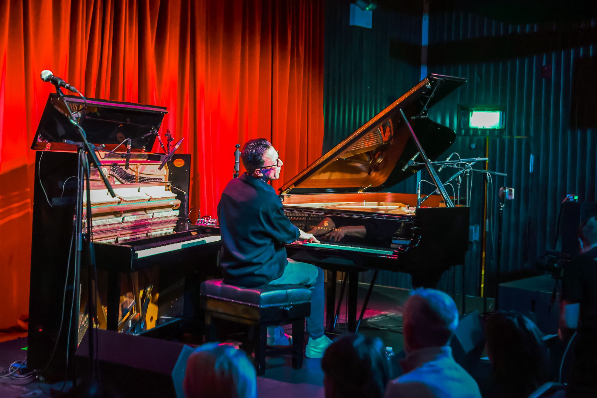 Wide view of the Sugar Club stage: Bill Laurance between the open upright and the concert grand, red curtain behind, the audience in silhouette