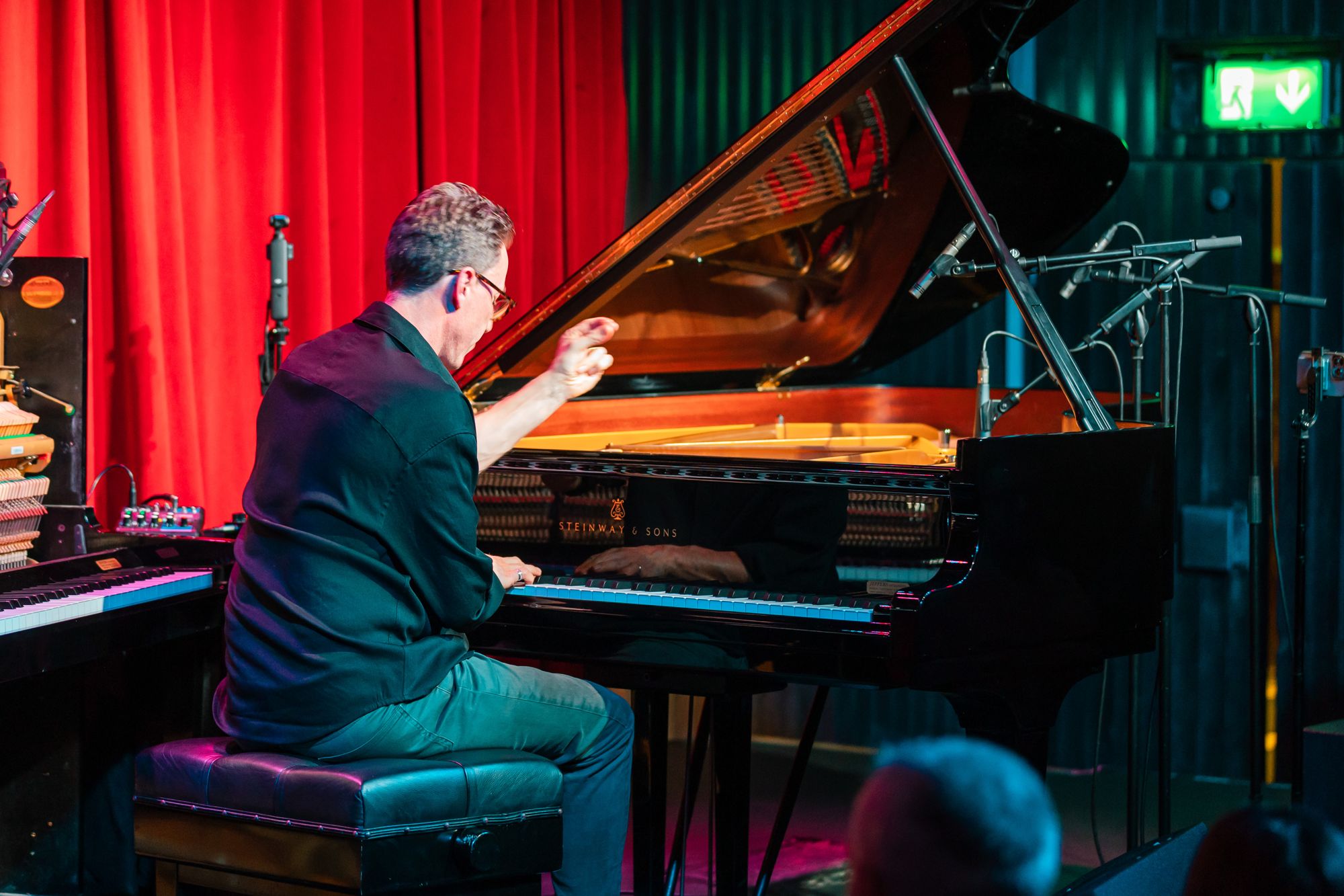 Bill Laurance seen from behind, one hand raised over the grand piano, red curtain to his left