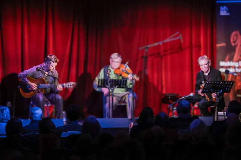 The musicians seated in a row on stage, red curtain behind