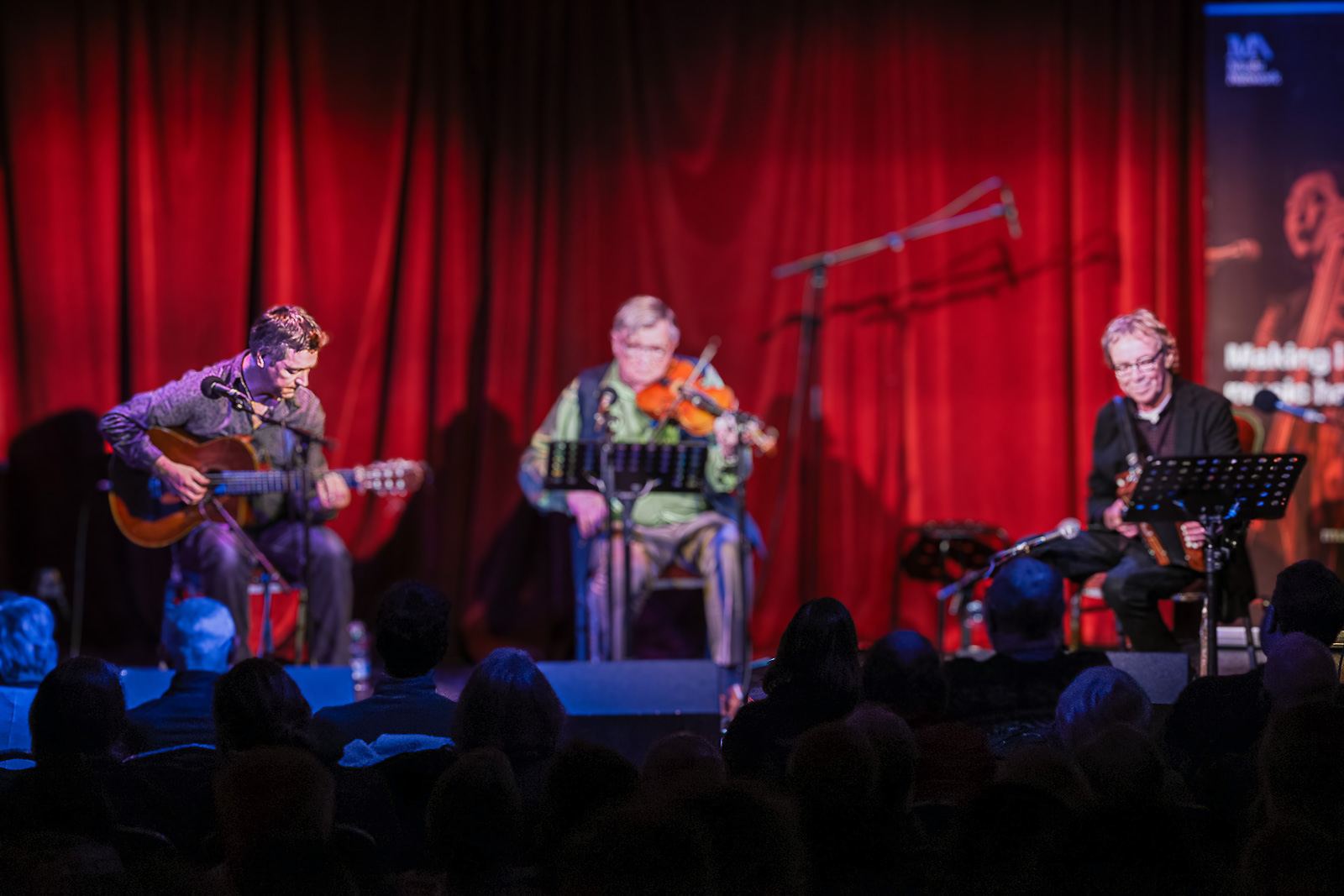 The musicians seated in a row on stage, red curtain behind