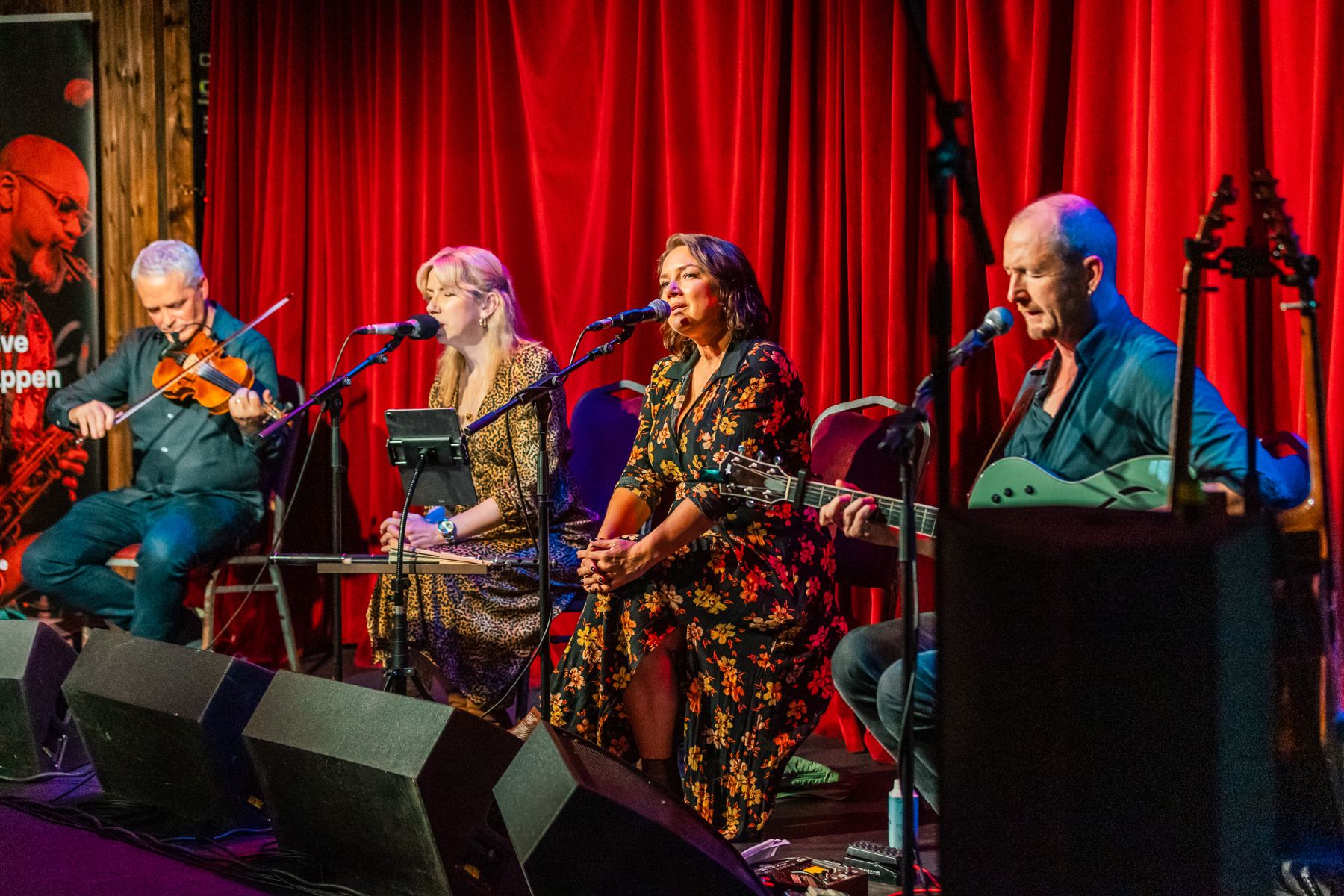 The four musicians seated across the stage of The Sugar Club in front of the red curtain