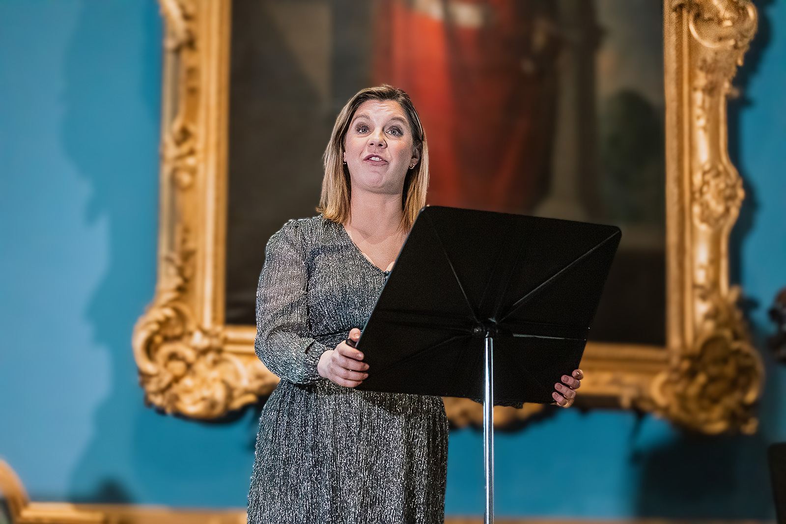 Alison Langer singing against the blue wall and gilded frames